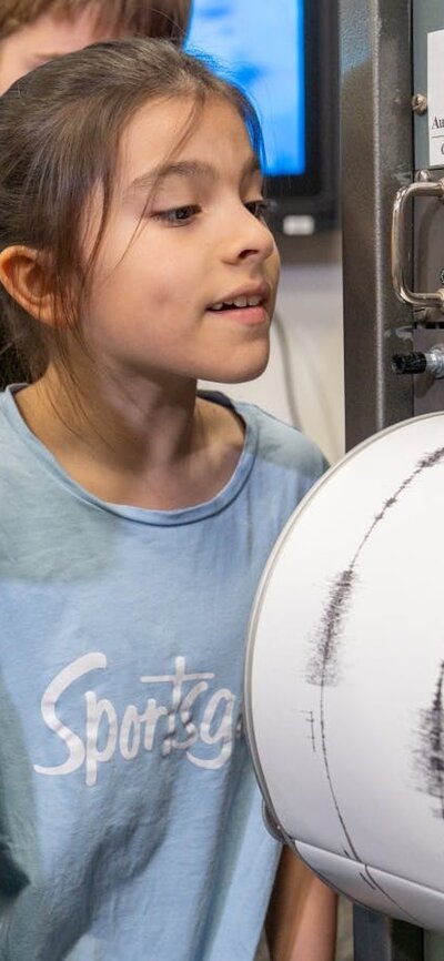 Two pre-teen girls look at a cylindrical drum that has white paper with black wave signals on it.
