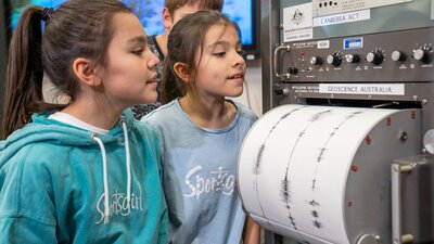 Two pre-teen girls look at a cylindrical drum that has white paper with black wave signals on it.