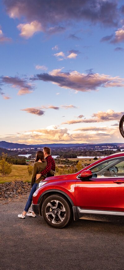 Two young kids on bikes and a couple lean against a red car bonnet overlooking the view of Black Mountain Tower at the National Arboretum Canberra