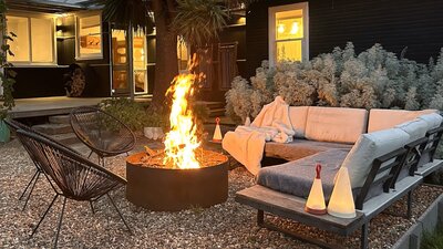 Firepit at dusk with outdoor couch, house in the background with lights on.