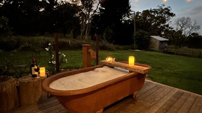 Rust-coloured bathtub at twilight on timber deck with bubblebath, candles, wine, string of lights