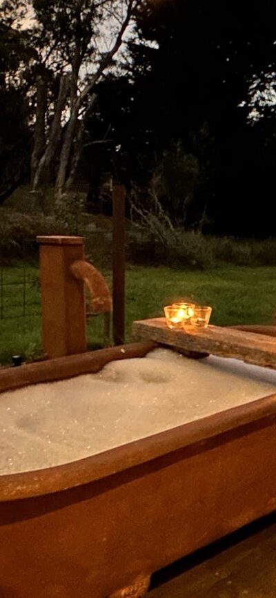 Rust-coloured bathtub at twilight on timber deck with bubblebath, candles, wine, string of lights