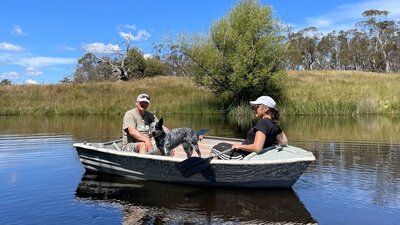 Man, woman & dog in small rowboat on water, blue sky, grass & trees behind