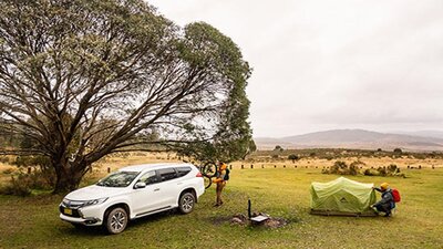 A man sets up a tent next to a fire ring and car at Long Plain Hut campground, Kosciuszko National
