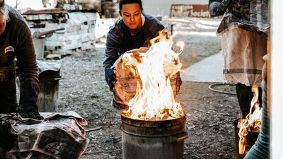 A female student reaches a metal garbage can lid towards a bin full of flames as part of raku firing