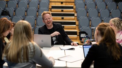 A man, laughing, sits at a table with a group of teenagers with laptops and notebooks in a theatre.