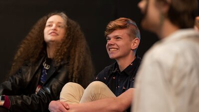 Three teenagers sit together in a theatre workshop, smiling as they enjoy watching a performance.