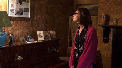 Dark living room at Mugga depicting lady lookin at old photos in wall and piano top