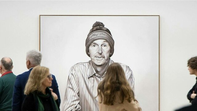 Visitors looking at Michael Peck's Play on (A portrait of Neale Daniher) 2025 © Michael Peck People stand in front of an oil painting of former Australian rules football player Neale Daniher.