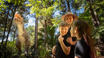 A mother and two children smile in awe at the size of the animatronic T-Rex  in lush fern gardens.