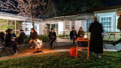 a group of people seated around a fire in a garden, outside a gallery.