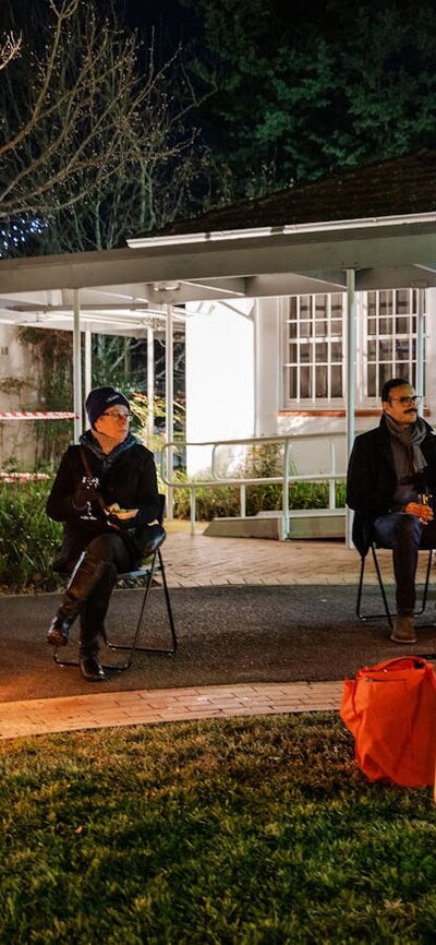 a group of people seated around a fire in a garden, outside a gallery.