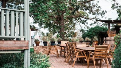Timber tables and chairs arranged on a paved area underneath a large tree.
