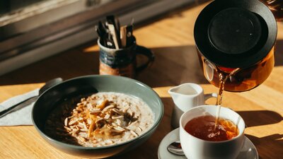A bowl of porridge sits on a table as a cup of tea is being poured from a jug.