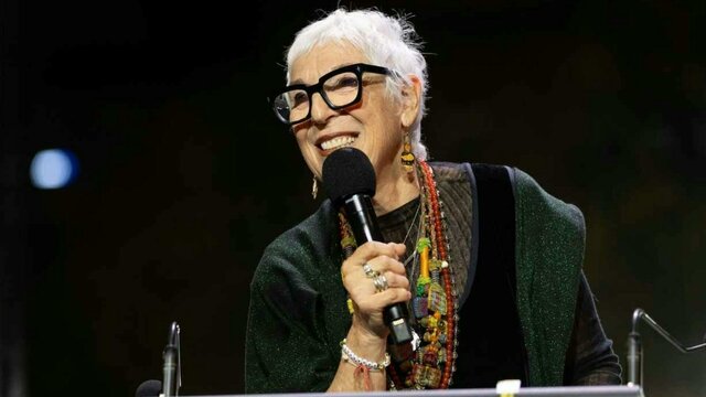 Ronni Kahn AO A woman with short grey hair and glasses holding a microphone on stage