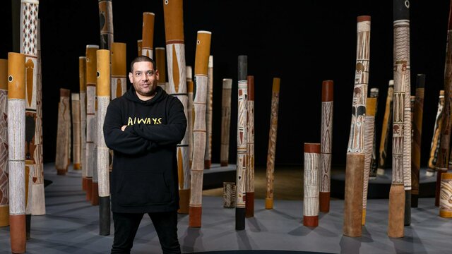 Tony Albert (Girramay/Yidinji/Kuku-Yalanji peoples) in The Aboriginal Memorial, 1987–88 A man standing in a gallery space with indigenous poles