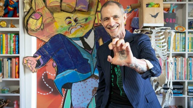 Andy Griffiths in his studio A man in a striped suit smiles and holds up his hand, which has a face tattooed in the palm.