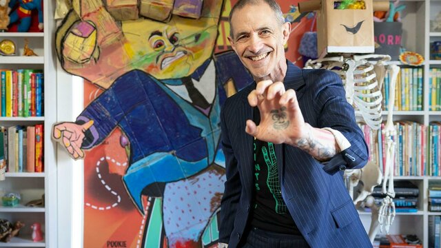 Andy Griffiths in his studio A man in a striped suit smiles and holds up his hand, which has a face tattooed in the palm.
