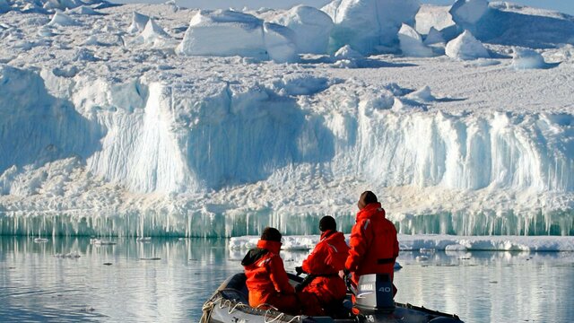 Expeditioners in an inflatable boat. Christopher Wilson / AAD Three people in a small inflatable boat looking at the large ice formation in front of them.