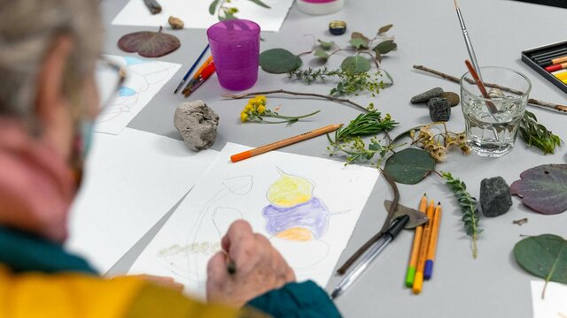 Art with Friends, 2022. National Gallery of Australia, Kamberri/Canberra. A photograph over the shoulder of an elderly person drawing gum leaves from real ones on the table