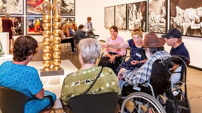 A group of people, some in wheelchairs, sit around a sculpture listening to a guide