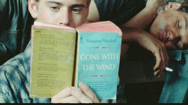 The Outsiders: The Complete Novel One male lying on couch sleeping, another sitting in front reading a book