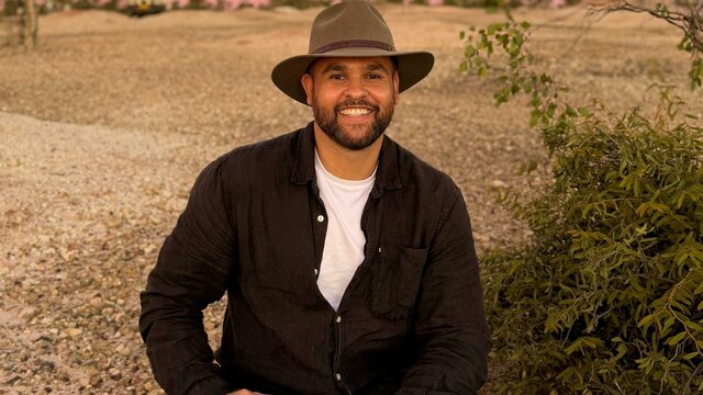 James Saunders A smiling man with a beard and a wide-brimmed hat sits on a gravelly path in a dry, bushy landscape.