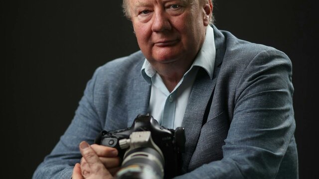 Mike Bowers A man with grey hair wearing a blue shirt and blazer, holding a large black camera.