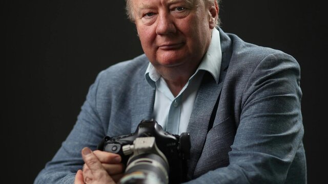 Mike Bowers A man with grey hair wearing a blue shirt and blazer, holding a large black camera.