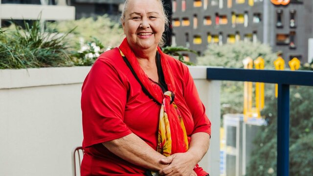 Isabel Goolagong A smiling woman sits on a balcony, wearing a red shirt and a red and yellow scarf.