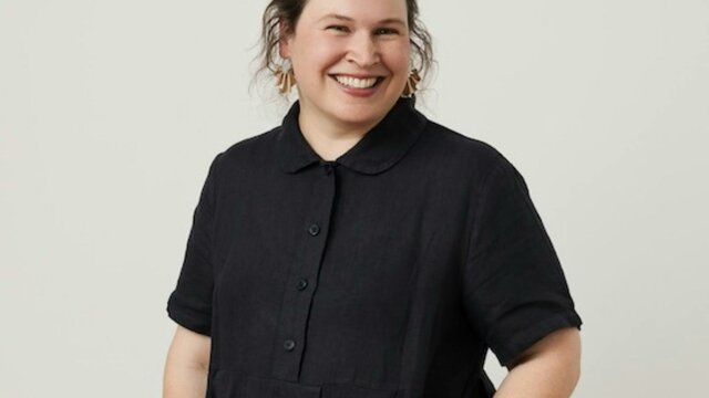 Catie Bradbear A smiling woman with dark hair in a bun, wearing a black short-sleeved button-up dress.