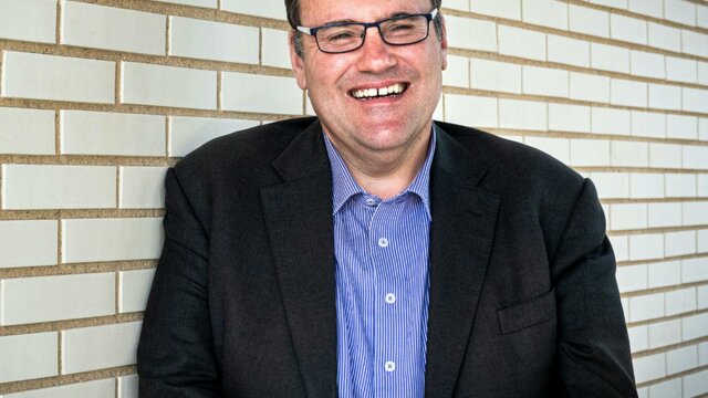 Frank Bongiorno AM A man with glasses wearing a sttriped button down shirt and black blazer, standing in front of a whi