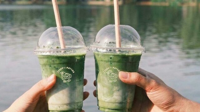 two takeaway matcha cups two matcha cups being held in two different hands doing a cheers in front of a lake background
