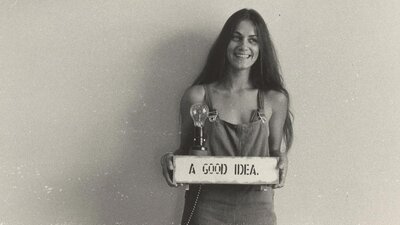 A young woman with long hair holding a sign that says 'A Good Idea' with a lightbulb on top of it.