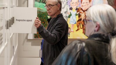 Man reading an interactive exhibition panel in front of Wizard of Oz political cartoon