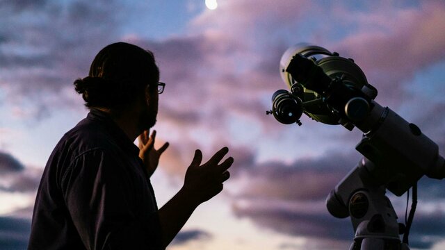 Man looks towards the moon with setting sun next to telescope