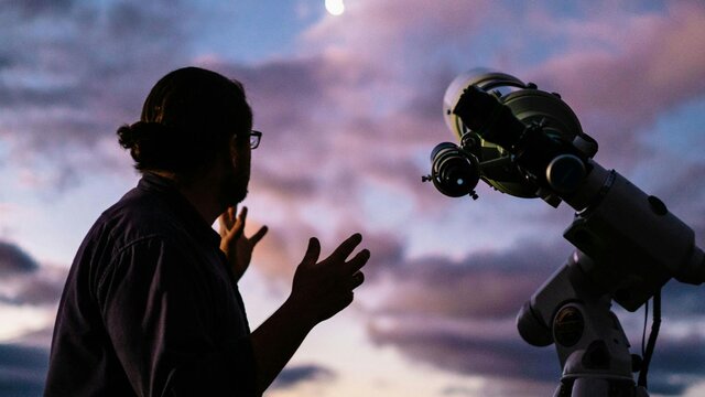 Man looks towards the moon with setting sun next to telescope