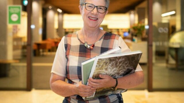 Dr Marie-Louise Ayres FAHA A smiling woman wearing glasses is pictured in a Library holding books
