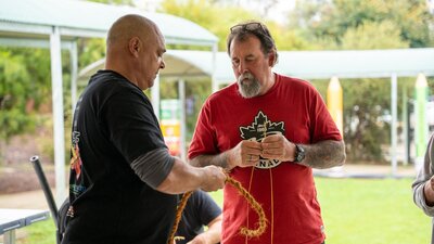 An Aboriginal man in a black tshirt showing another man how to make rope using traditional methods.