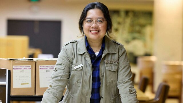 Dr Pauline Basilia A woman in a green jacket stands leaning on a table covered with papers