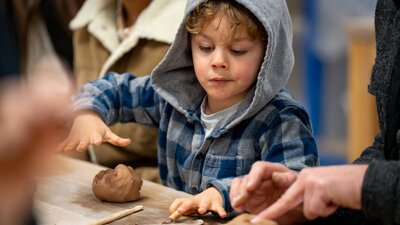 A young boy wearing a blue and grey hoodie makes a piece of art out of brown clay.