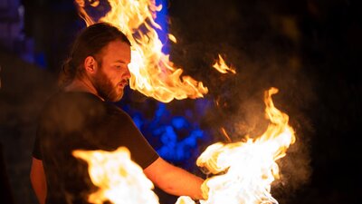 A young man with a black beard and t-shirt is surrounded by fire which he twirls on a stick