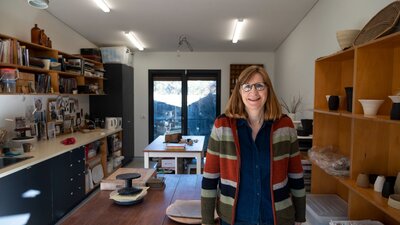 A lady with shoulder length red hair and glasses stands in her pottery studio, smiling at the camera