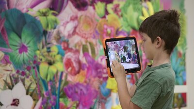 A boy holding an orange iPad recording a wall with a floral pattern