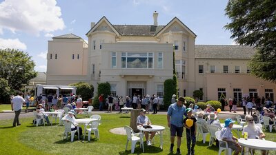 people sit at tables and chairs on lawn with a three storey white house in the background