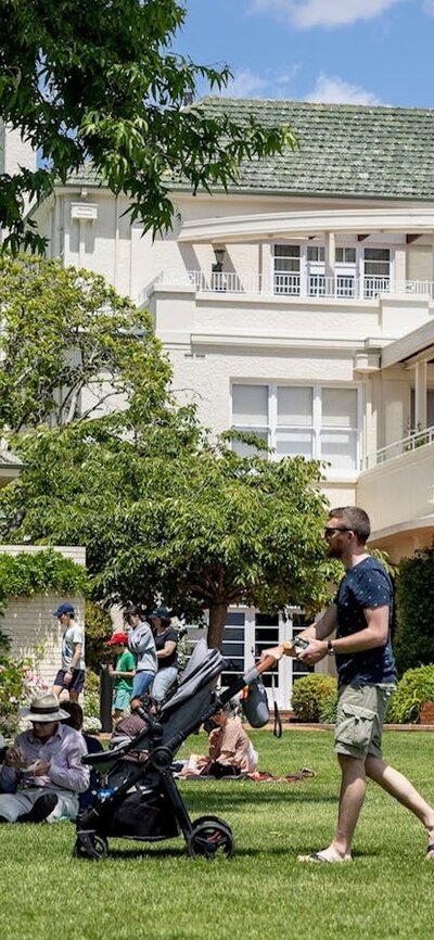 People sitting and standing on lawn with a large white house with a green roof in the background