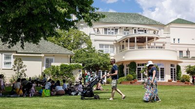 People sitting and standing on lawn with a large white house with a green roof in the background