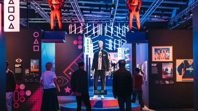 Hallyu! The Korean Wave exhibition Exhibit with mannequins in red jumpsuits, surrounded by neon lights and displays.