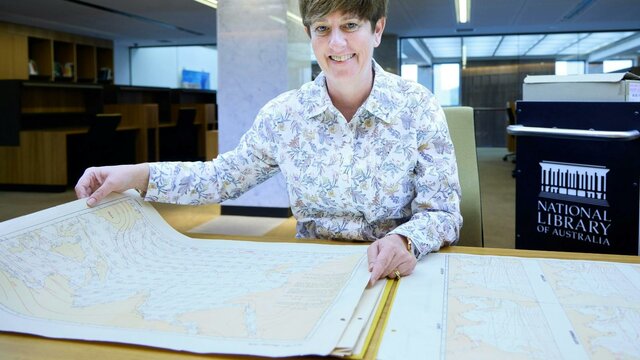 Dr Margaret Cook A smiling woman in a floral shirt sits at a desk, holding open a large historical map.