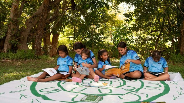 Image courtesy of the Indigenous Literacy Foundation. A group of diverse young children are sitting outdoors under lush green trees, engaged in reading.